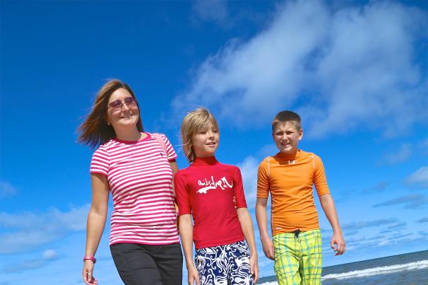 A woman and two young children walk along the beach. They are dressed in bright colored clothing. Water and a blue sky can be seen in the background.