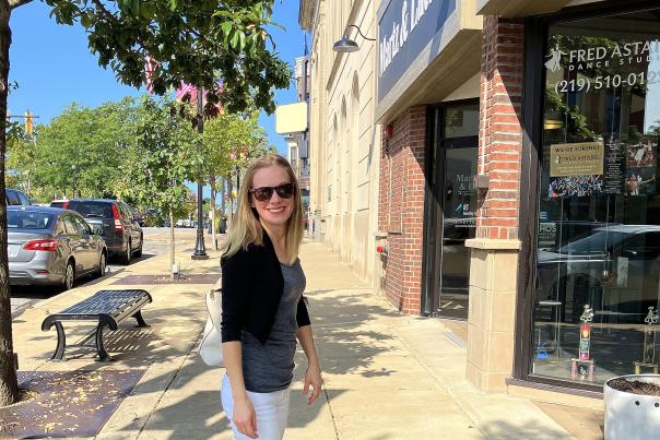 A woman with sunglasses stands on a sidewalk in front of storefronts.