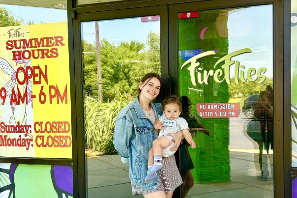 A smiling woman in a denim jacket holds a baby outside the entrance of a store called Fireflies.