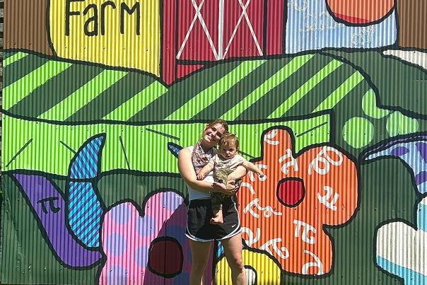 A woman holding a child stands in front of a mural at Sunset Hill County Park
