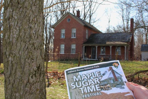 A hand holds a "Maple Sugar Time Festival" program in front of a historic building