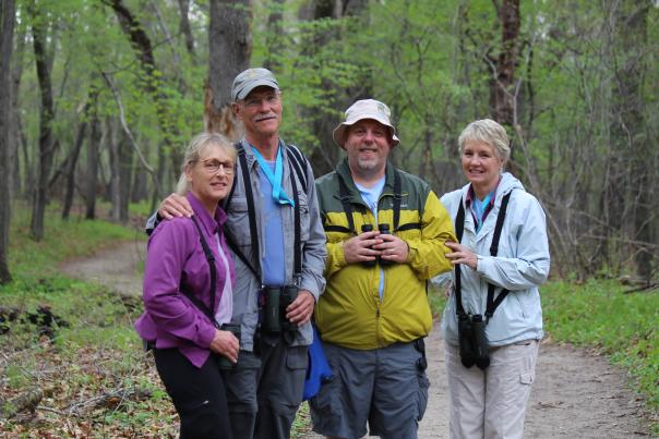 A group of four people stand together on a trail. They are looking at the camera, smiling. Woods are in the background