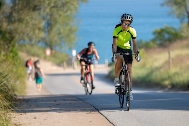 Bicyclists ride up hill on a paved road. A lake is in view in the background.