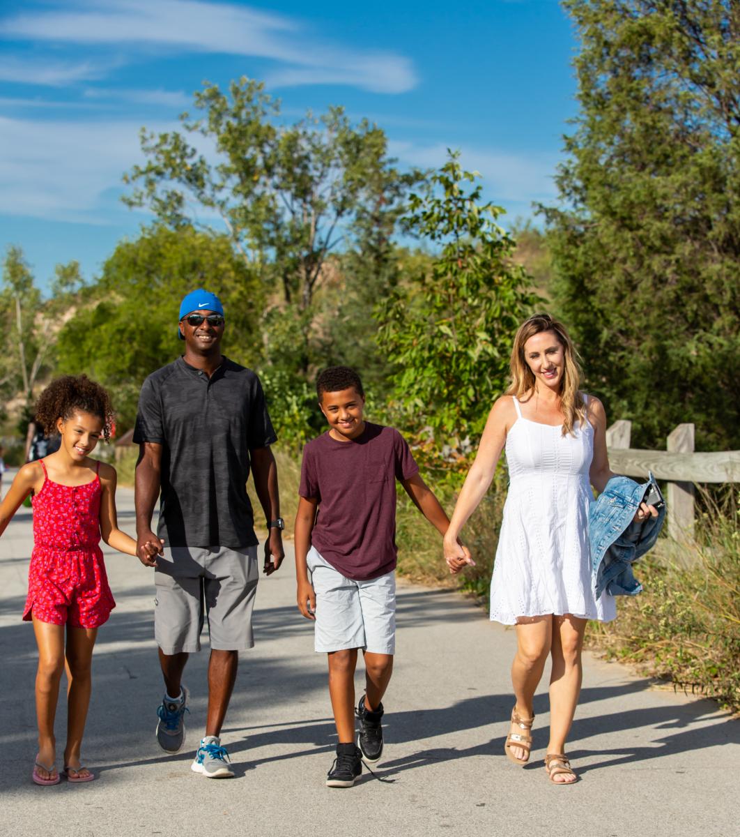 A family of four holds hands as they walk along a paved path through the dunes.