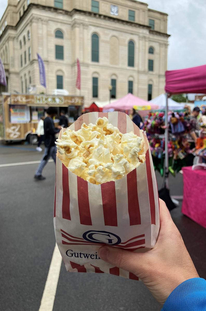 A red and white striped bag of popcorn is held up in front of a historical building.