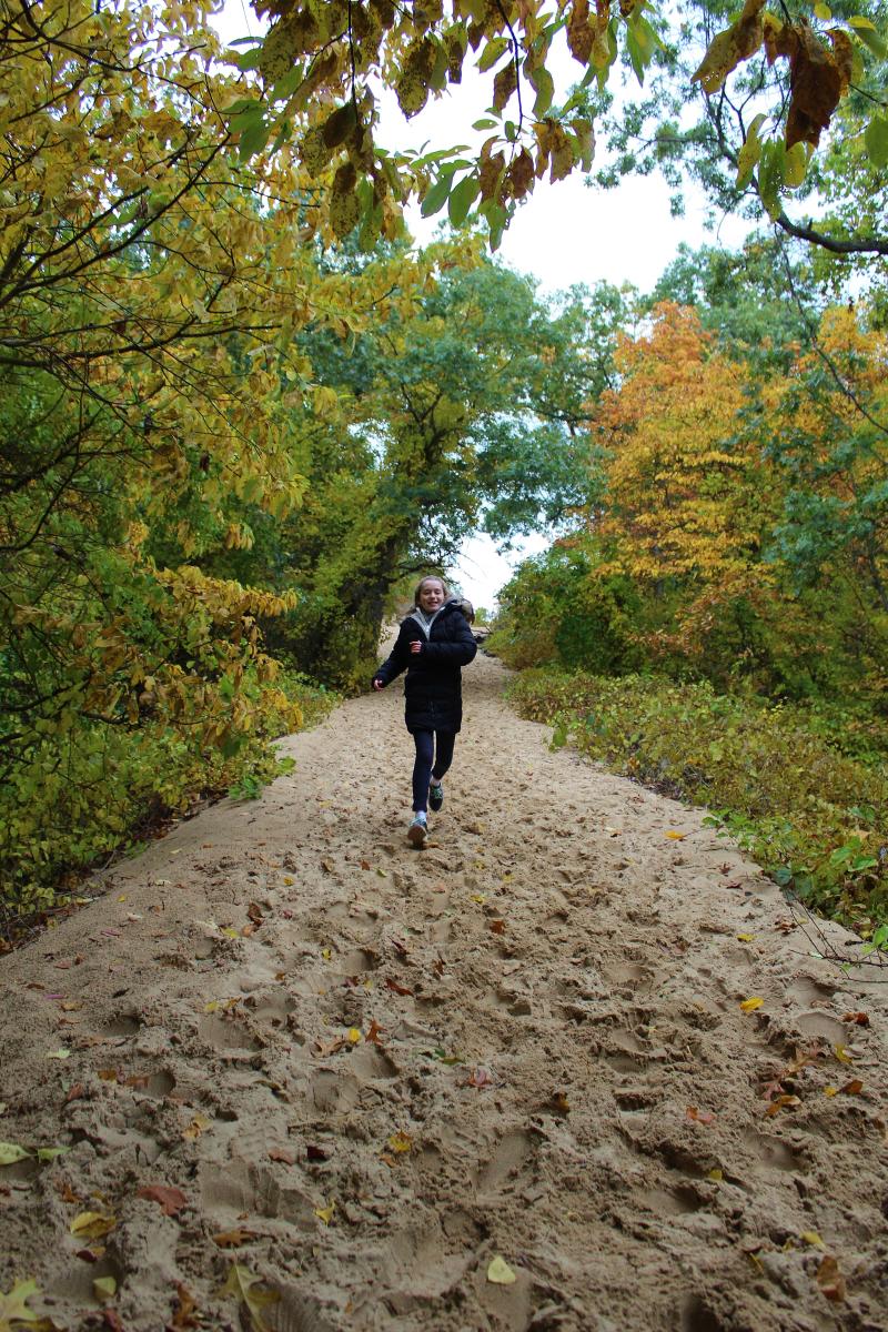 Girl runs up a steep sand dune. Fall trees line the sandy trail.