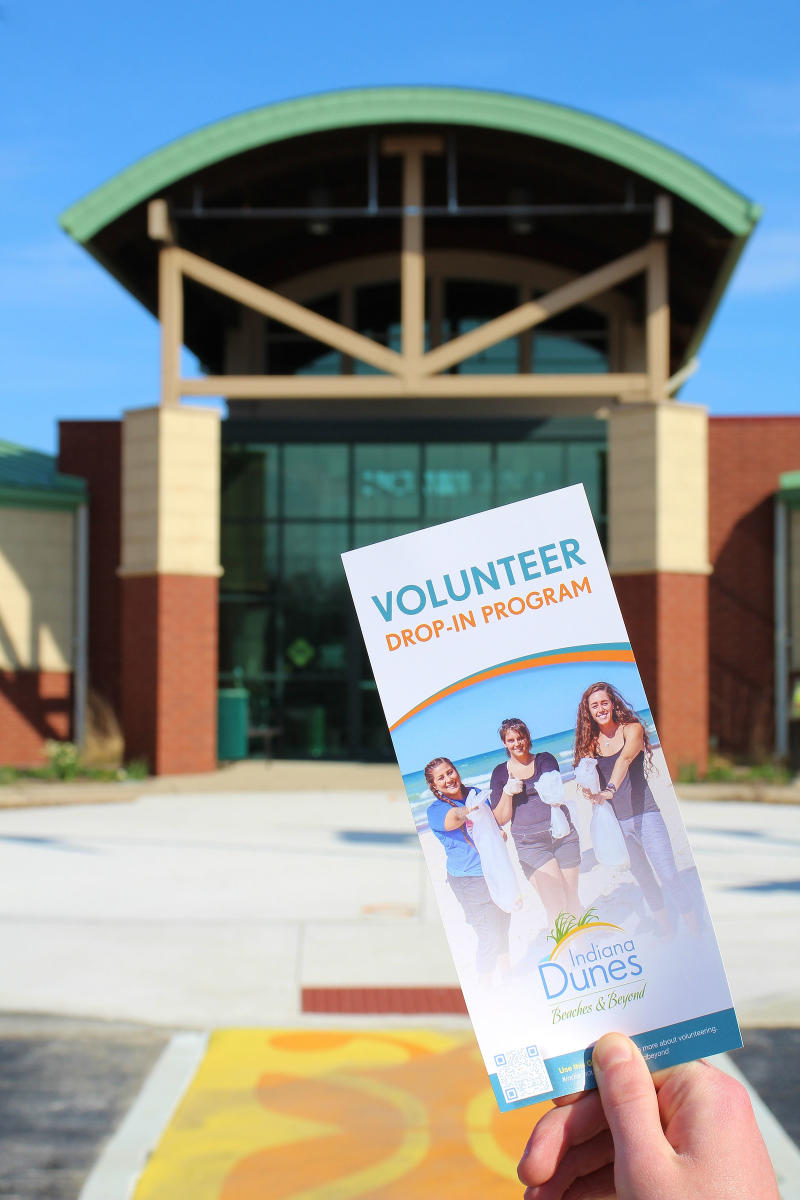 A hand holds a volunteer brochure in front of a brown and green building. A yellow and orange crosswalk is at the bottom.