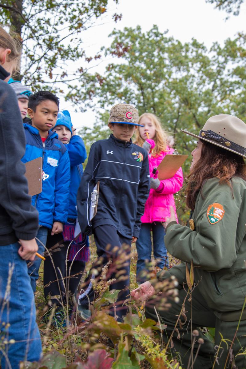 A group of school children listen to a National Park Ranger outdoors.