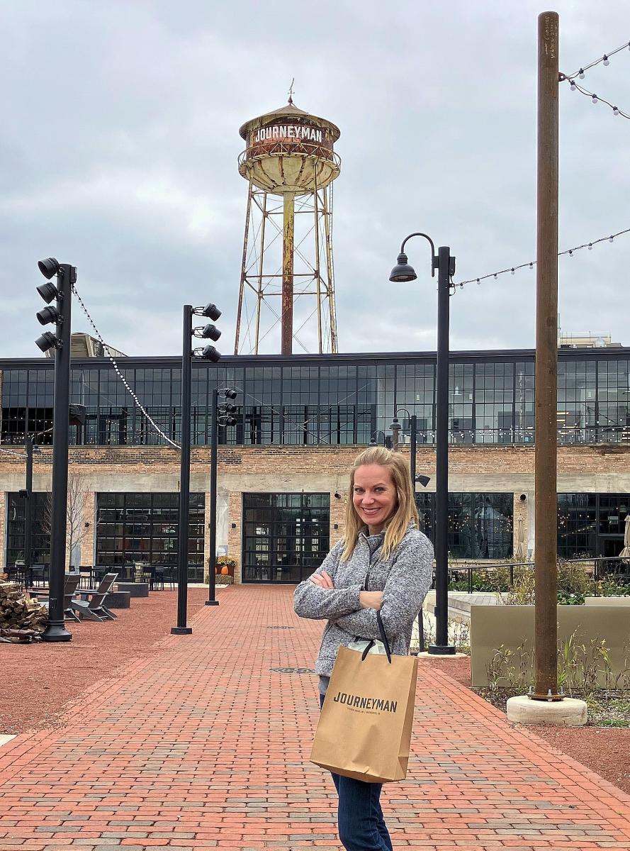 A woman in a grey sweater holds a brown shopping bag. She stands in front of a converted factory building with a water tower that says Journeyman