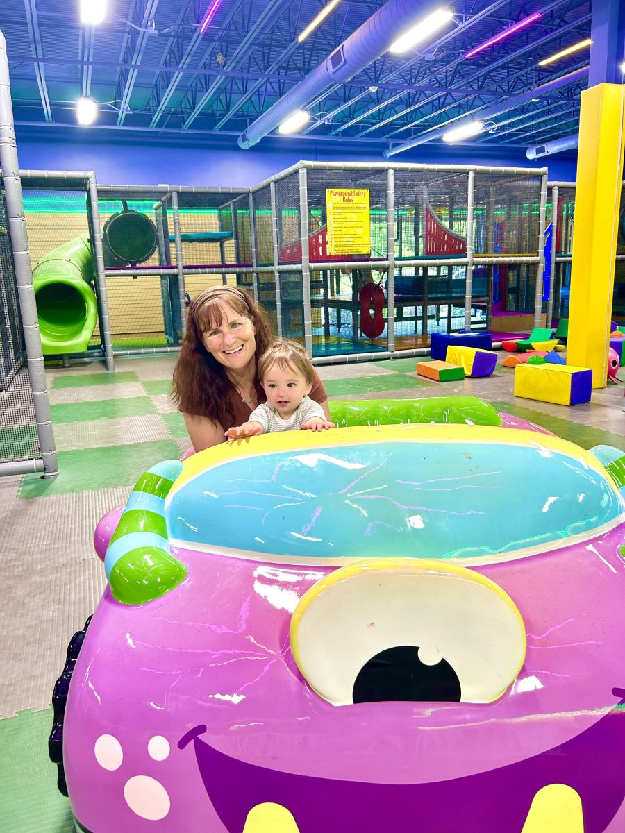 A woman and toddler smiling inside an indoor playground behind a large purple toy car.
