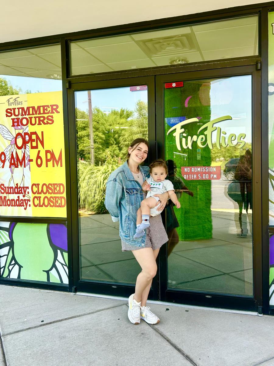 A smiling woman in a denim jacket holds a baby outside the entrance of a store called Fireflies.