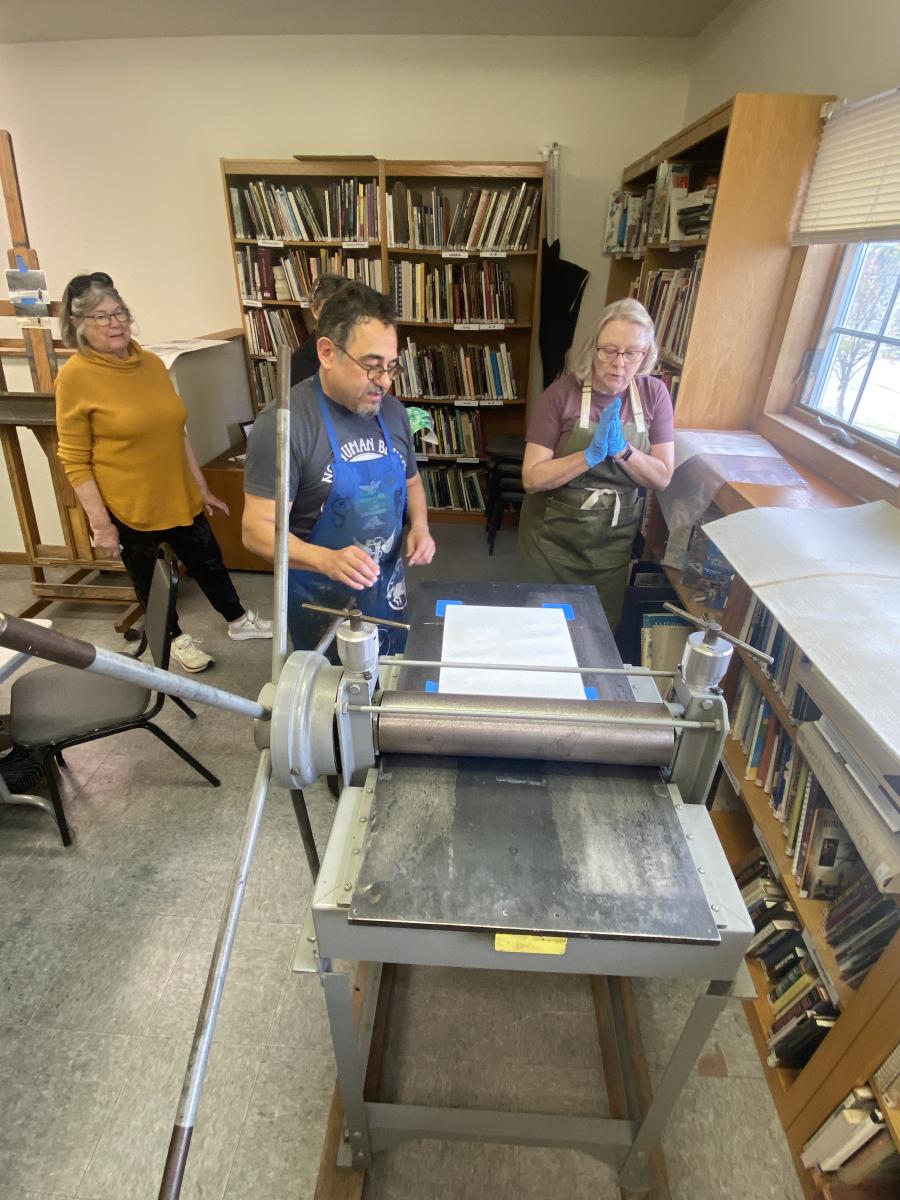 Three people stand around a printing press in a brightly lit studio filled with bookshelves.