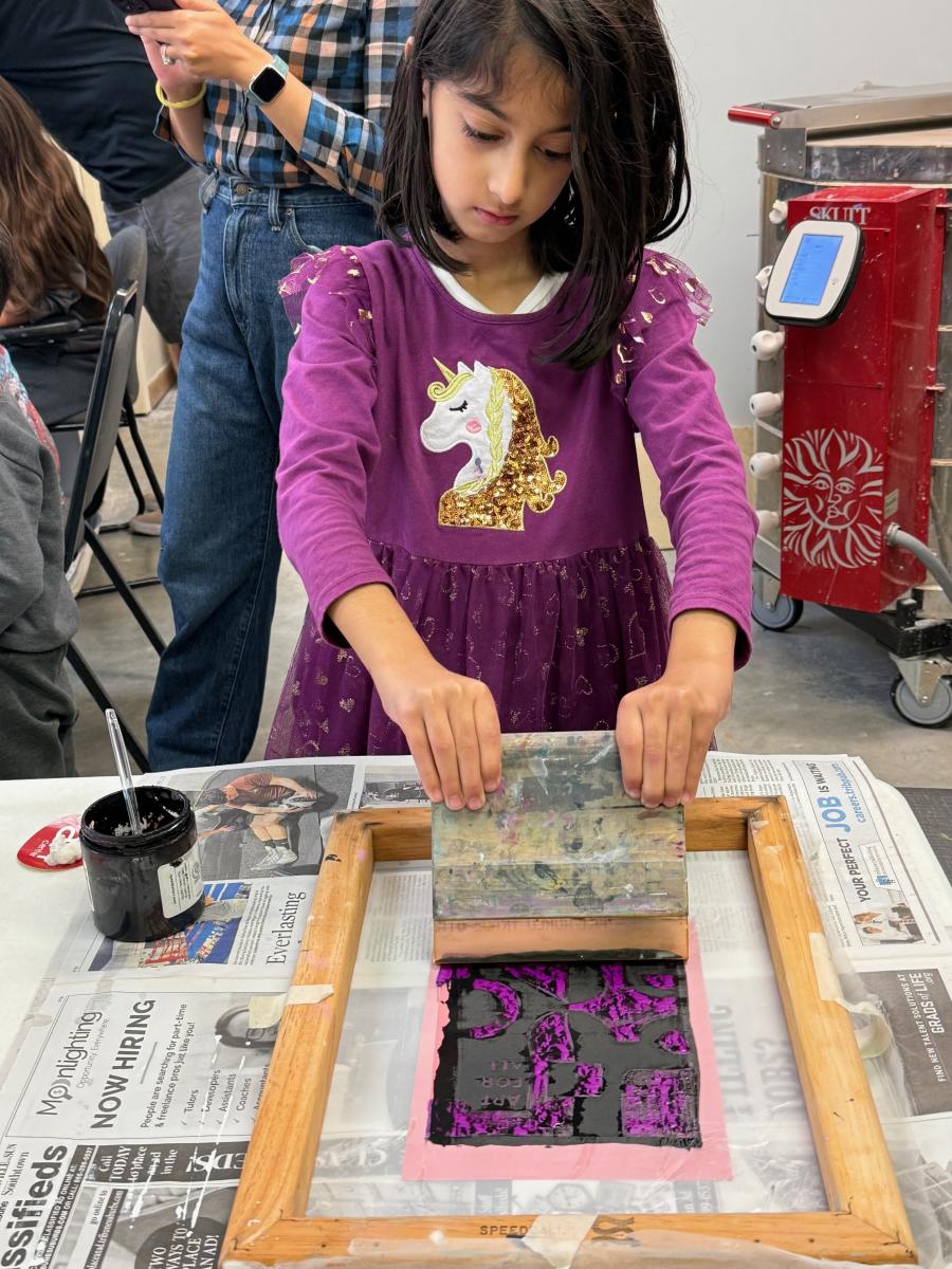 A young girl in a purple unicorn dress pulls a squeegee across a screen-printing frame over pink paper.
