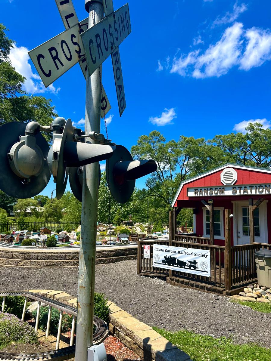 A miniature railroad crossing signal and a red "Ransom Station" building with a sign for the Illiana Garden Railroad Society.