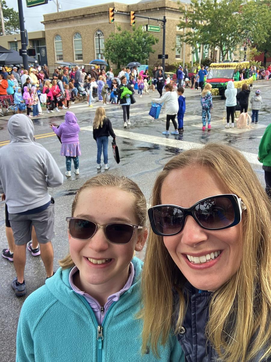 A mom and daughter wearing sunglasses look at the camera. A crowd lines the street behind them.