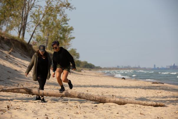 Two women are hiking along the beach. Trees are to the left. The water is to the right. One woman is stepping on a tree branch.