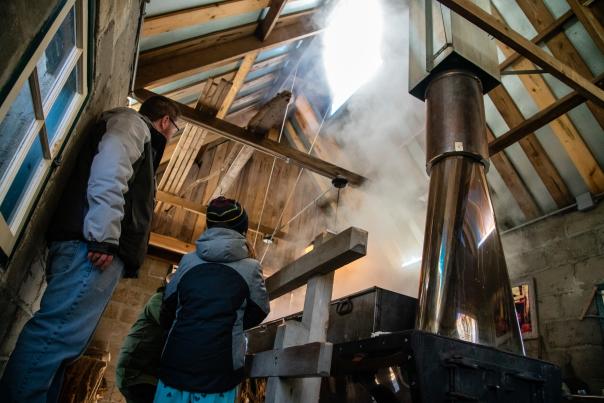 A cozy sugar shack with wooden walls and an open-beam ceiling fills with steam from a maple syrup evaporator. Two people, bundled in winter clothing, stand nearby, watching the process. A metal chimney pipe reflects the warm light, adding to the rustic atmosphere