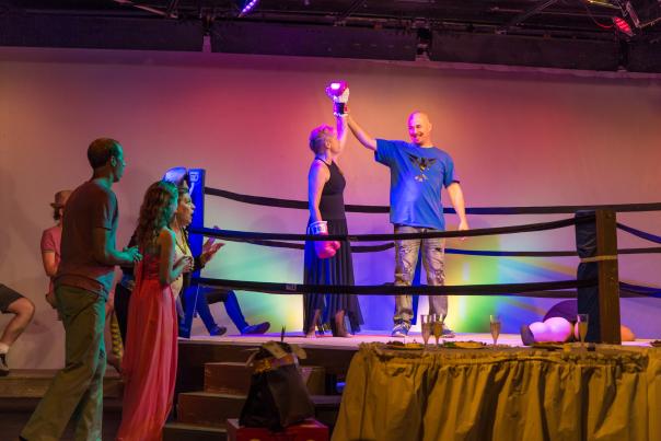 A man and woman stand in a boxing ring on a theatre stage. The woman is in a long black dress. The man is raising the woman's boxing gloved hand up in the air.