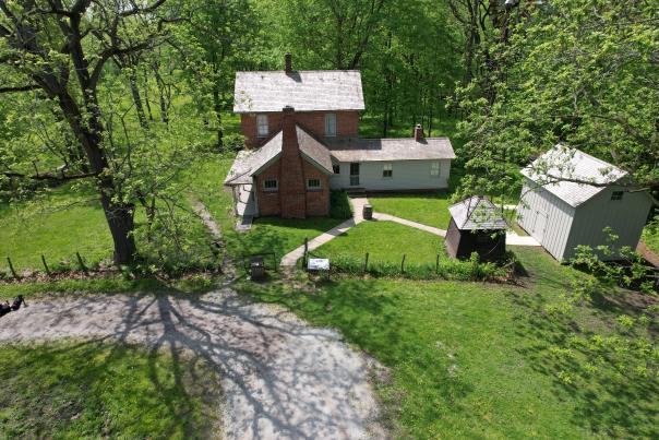 Aerial view of a historical building. A shed and windmill house sits to the left. A gravel road arches on the right bottom.