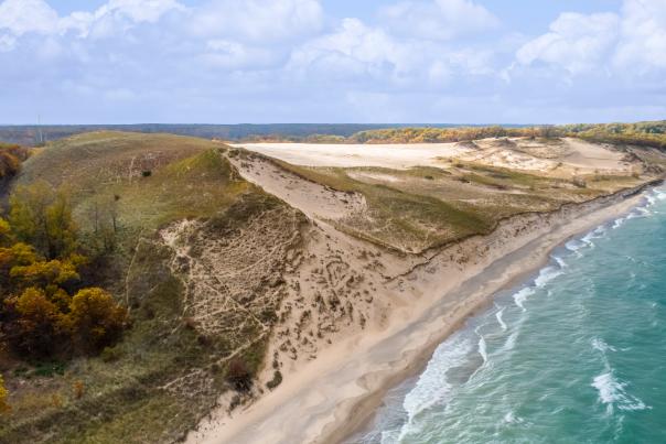 Aerial view of sandy dunes beside autumn bushes and a turquoise lake with gentle waves beneath a cloudy sky.