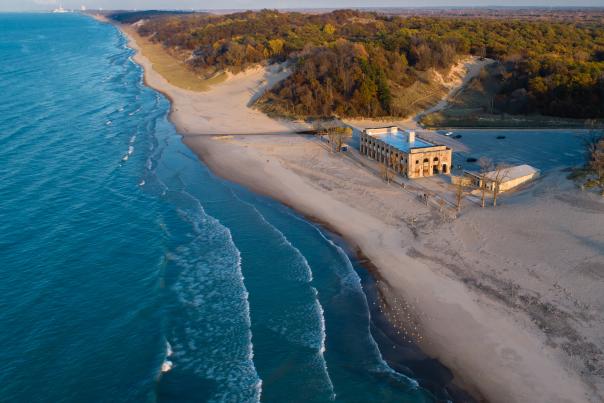 Bright blue water with white cresting waves near the shore sits to the left. On the right is a long sandy beach that spreads to the horizon line. Wooded dunes extend from the beach and into the distance. A stone building sits on the beach with a parking lot behind it.