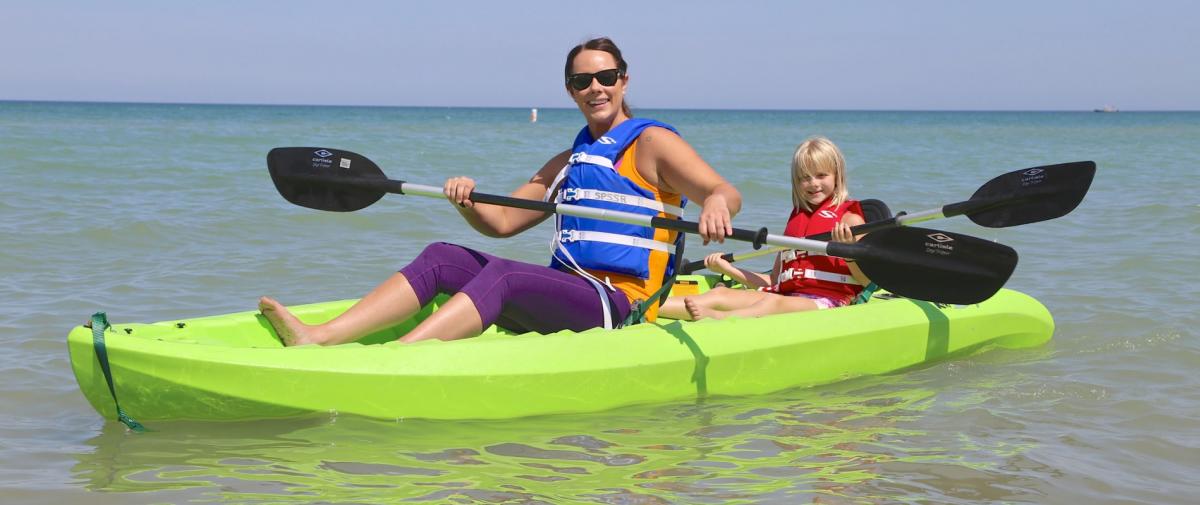 A woman and child sit on a double kayak on the water. Each has a lifejacket on and paddles in their hands.