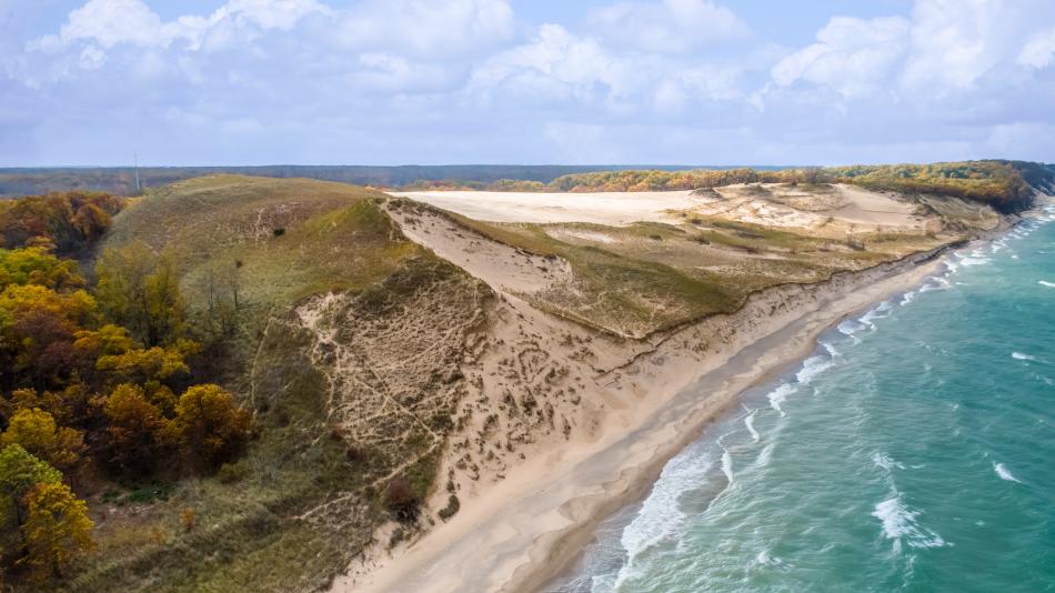 Aerial view of sandy dunes beside autumn bushes and a turquoise lake with gentle waves beneath a cloudy sky.