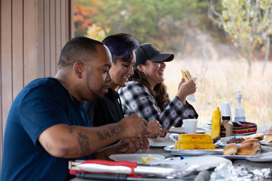 Three people sit at a picnic table. Plates of burgers, corn on the cob, and other food is on the table.