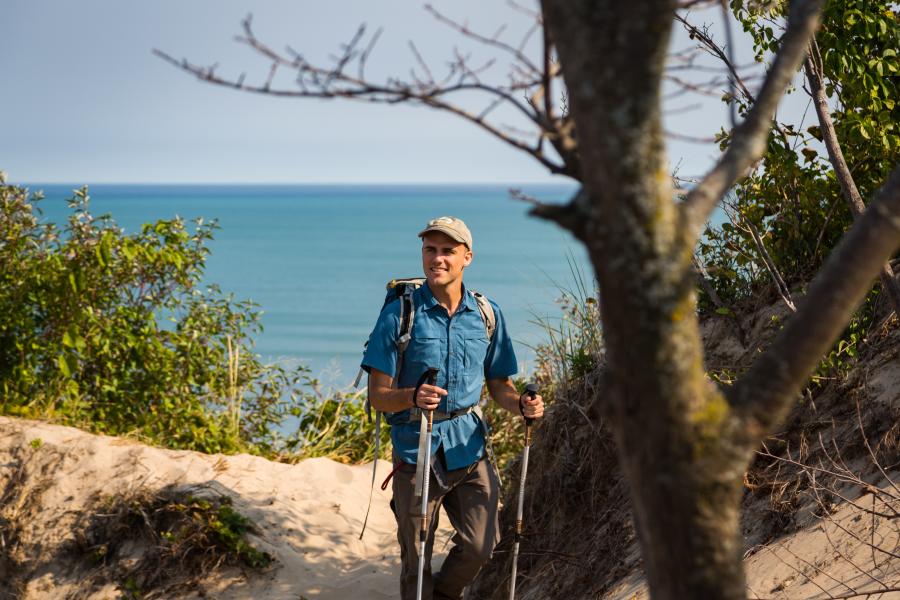 A man in a blue shirt and light cap hikes on a sandy trail with hiking poles. Lake Michigan can be seen in the background.