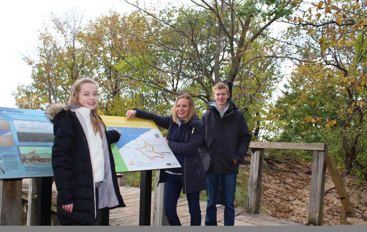 Three people in fall jackets pose next to a sign with trees in the background