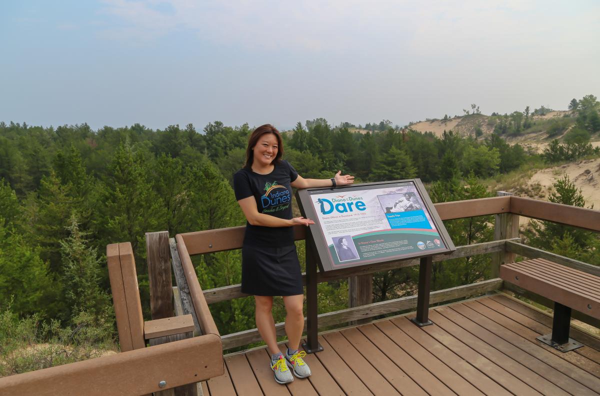 A woman in a shirt that says "Indiana Dunes Beaches & Beyond" stands on a deck next to a "Diana Dunes Dare" sign. In the background are tree-covered dunes.
