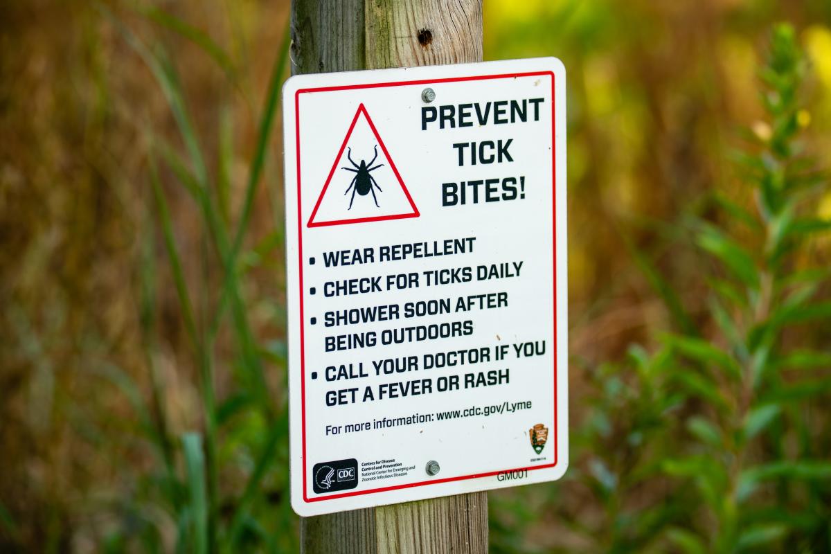 A white sign with a red border and a black tick icon warns to "PREVENT TICK BITES!" with prevention tips.