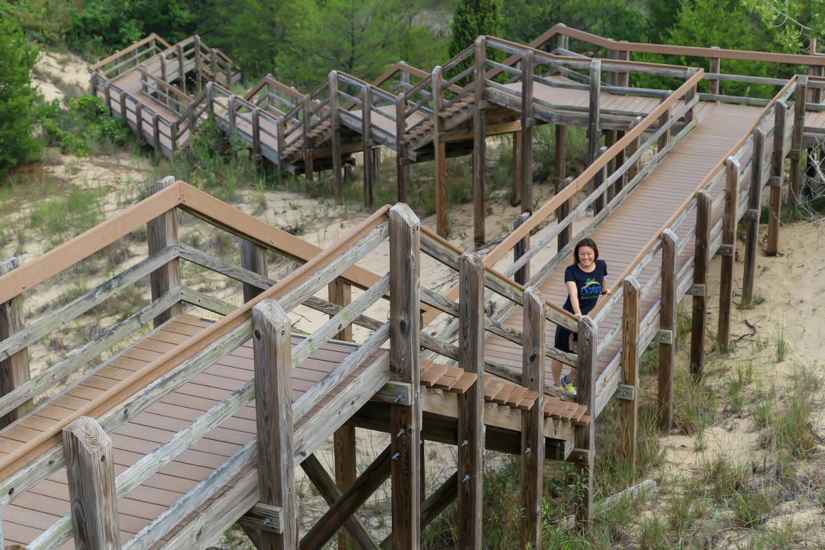 A woman wearing a black shirt stands on a wooden staircase and is surrounded by sand and vegetation.