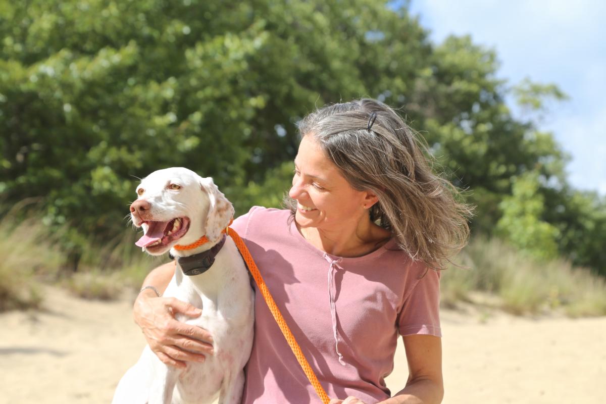 A woman with gray hair smiles at a white and brown spotted dog on a leash, with green trees in the background.