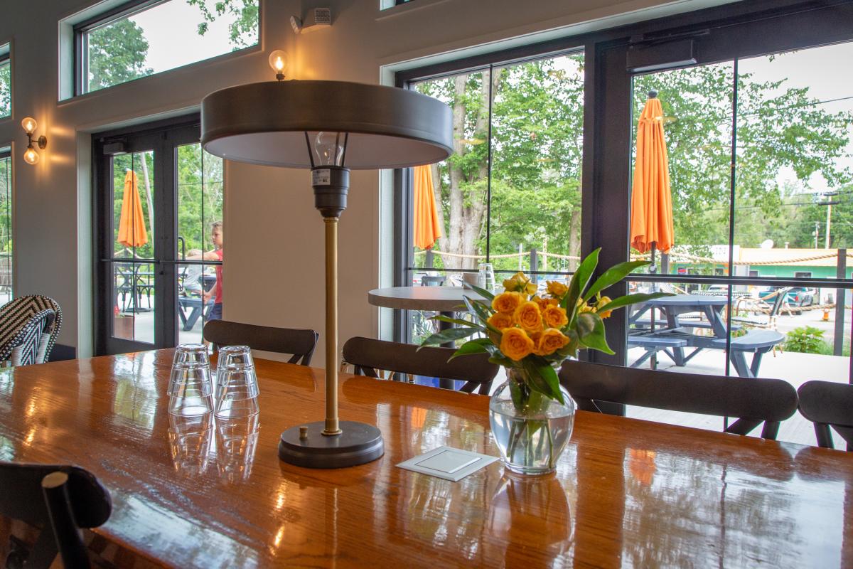 An indoor restaurant table with flower settings. Through the window, outdoor dining tables with orange umbrellas are seen.