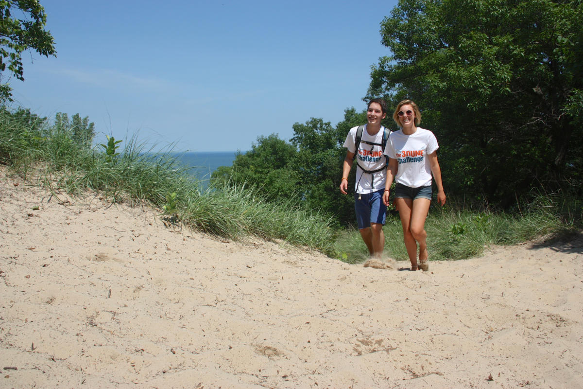Two young people hike on a sandy dune. Lake Michigan and foliage can be seen in the background.