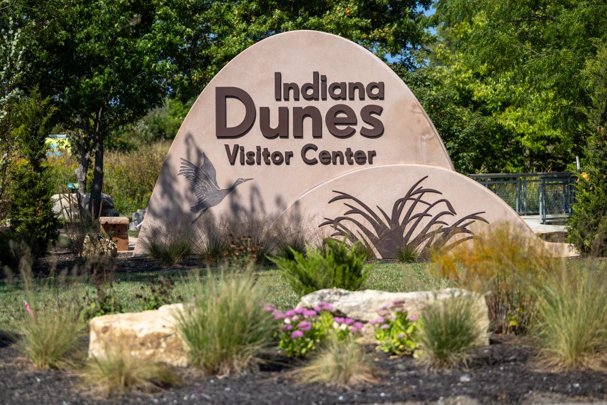 A dune-shaped sign says "Indiana Dunes Visitor Center" and has imagery of a sandhill crane and marram grass.