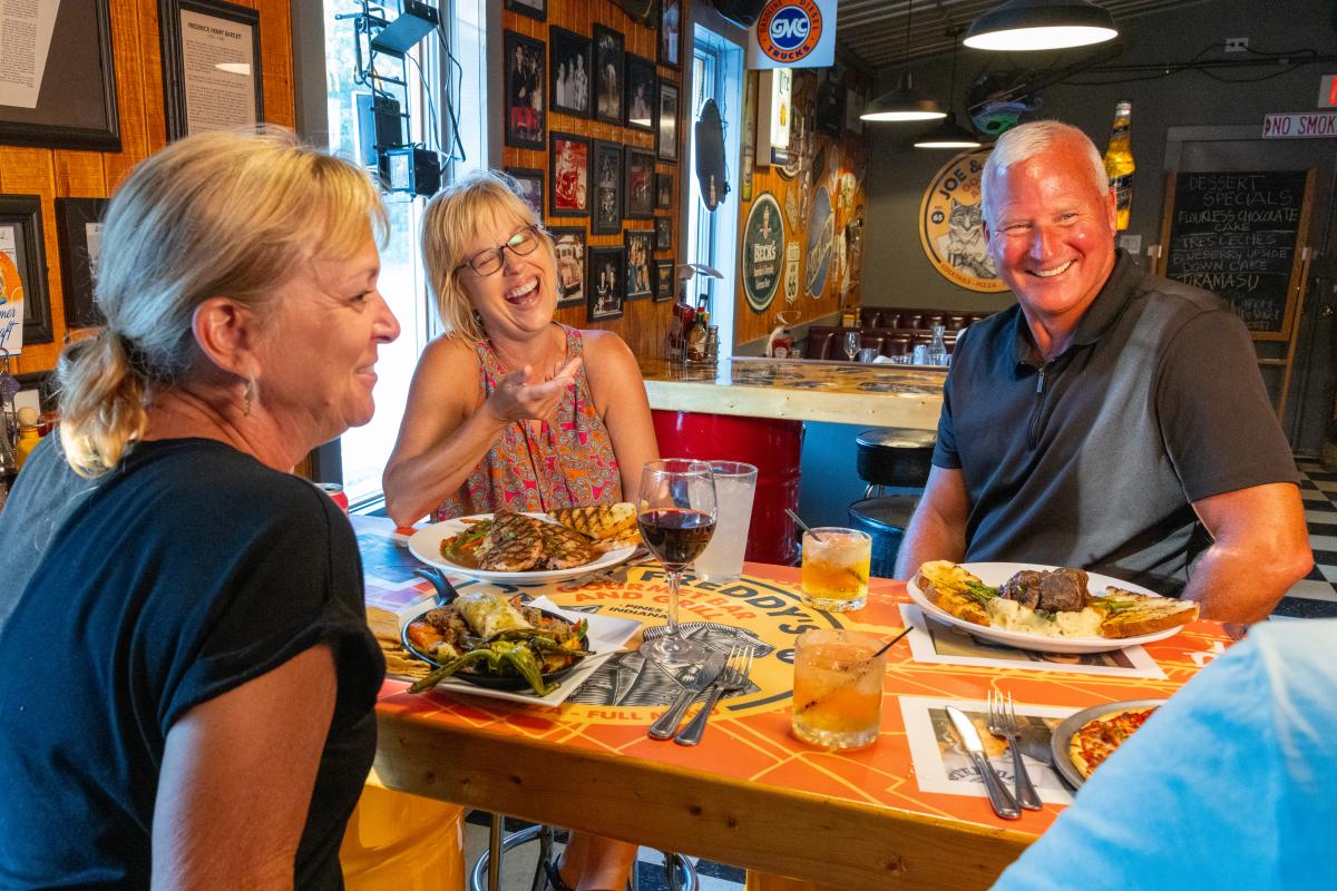 Three people smiling and enjoying a meal together at Joe and Freddy's restaurant, with food and drinks on the table and a warm, rustic bar interior in the background.