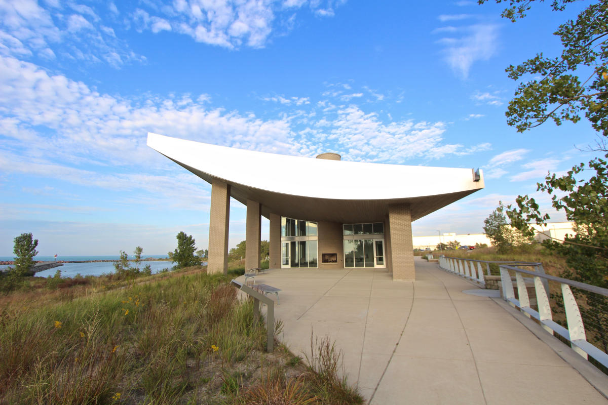 A building with a curved roof sits near the lake. The sky behind the building is bright blue with scattered clouds.