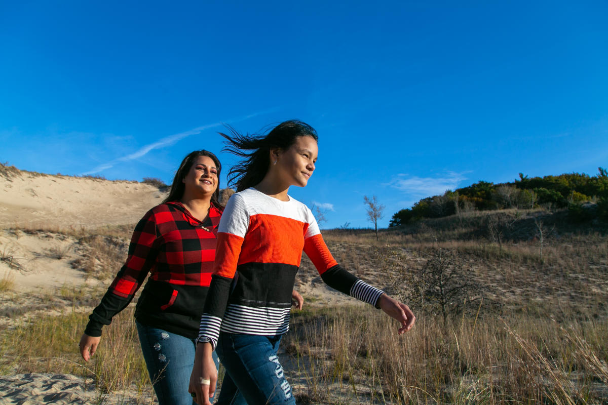 A woman and teen hike in the dunes. The bright blue sky is behind them. Vegetation on dunes covers the ground.