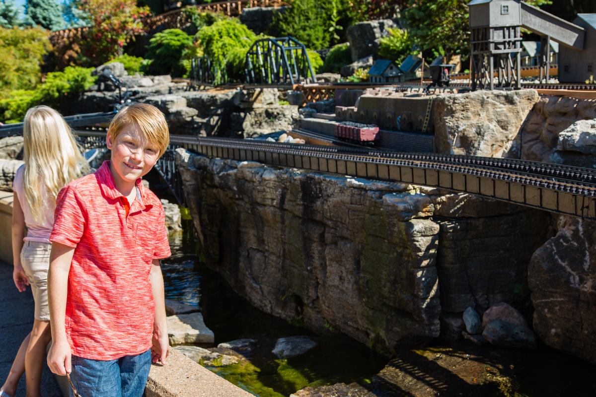 A young boy and girl stand by an outdoor miniature train display. The boy, wearing a red shirt, is looking at the camera.