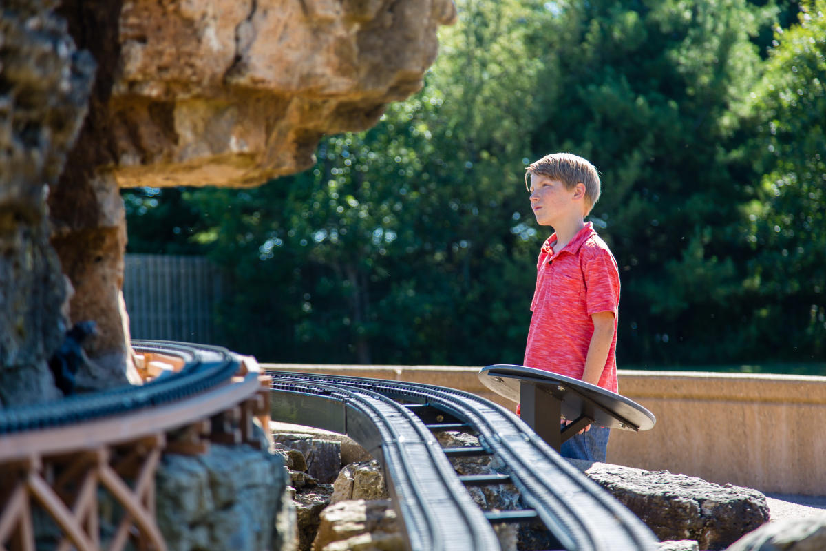 A boy wearing a red shirt looks at an outdoor train display. Tracks curve around a rocky landscape.