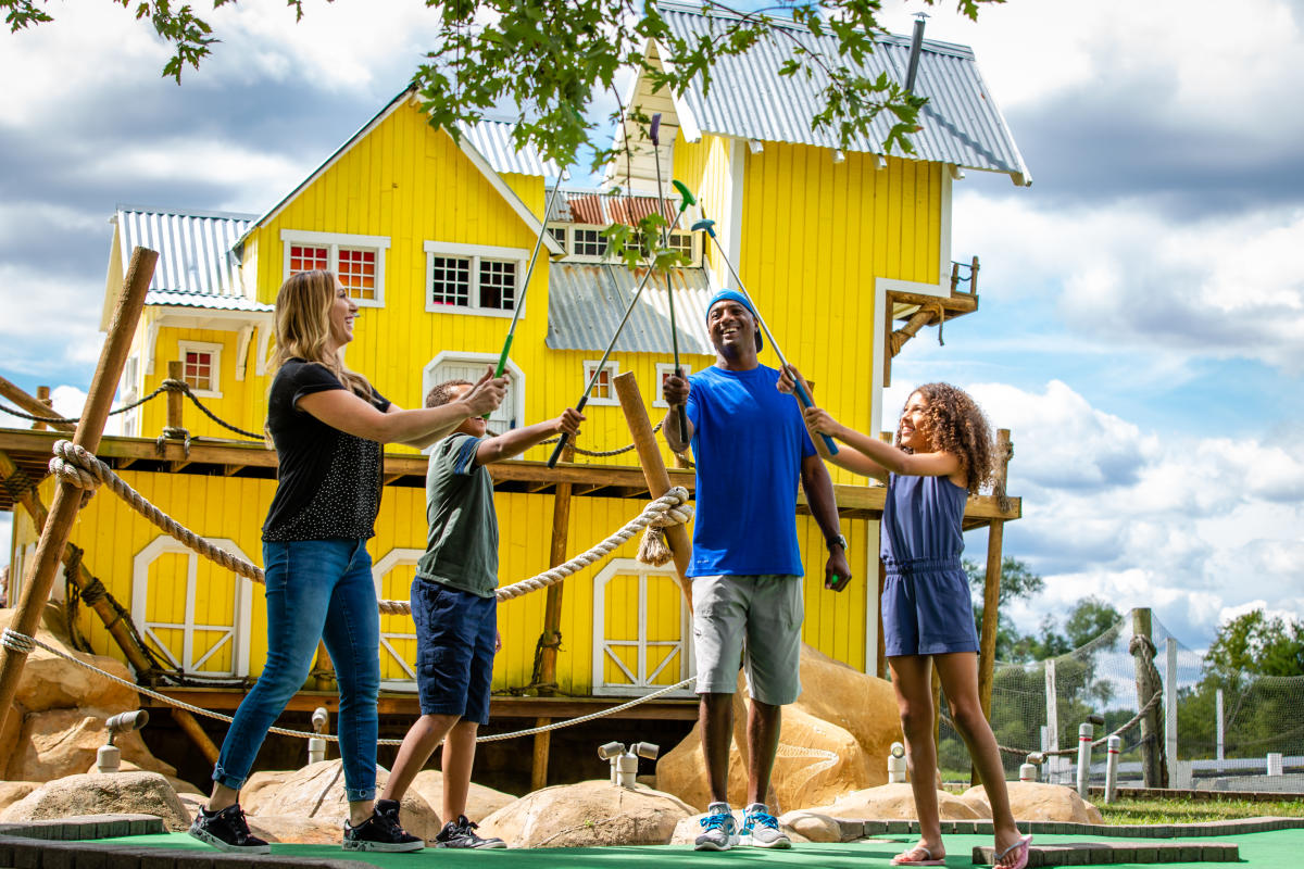 A family of four holds up golf clubs in celebration in front of a bright yellow house on a mini golf course.