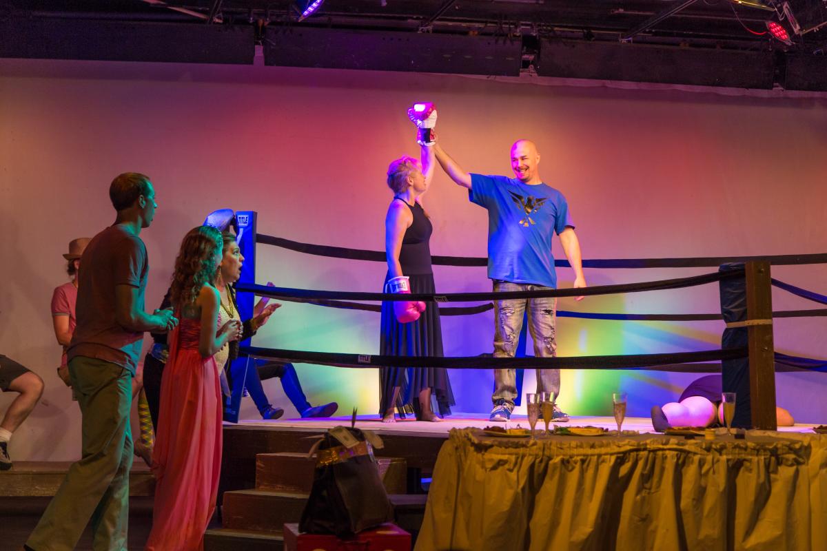 A man and woman stand in a boxing ring on a theatre stage. The woman is in a long black dress. The man is raising the woman's boxing gloved hand up in the air.