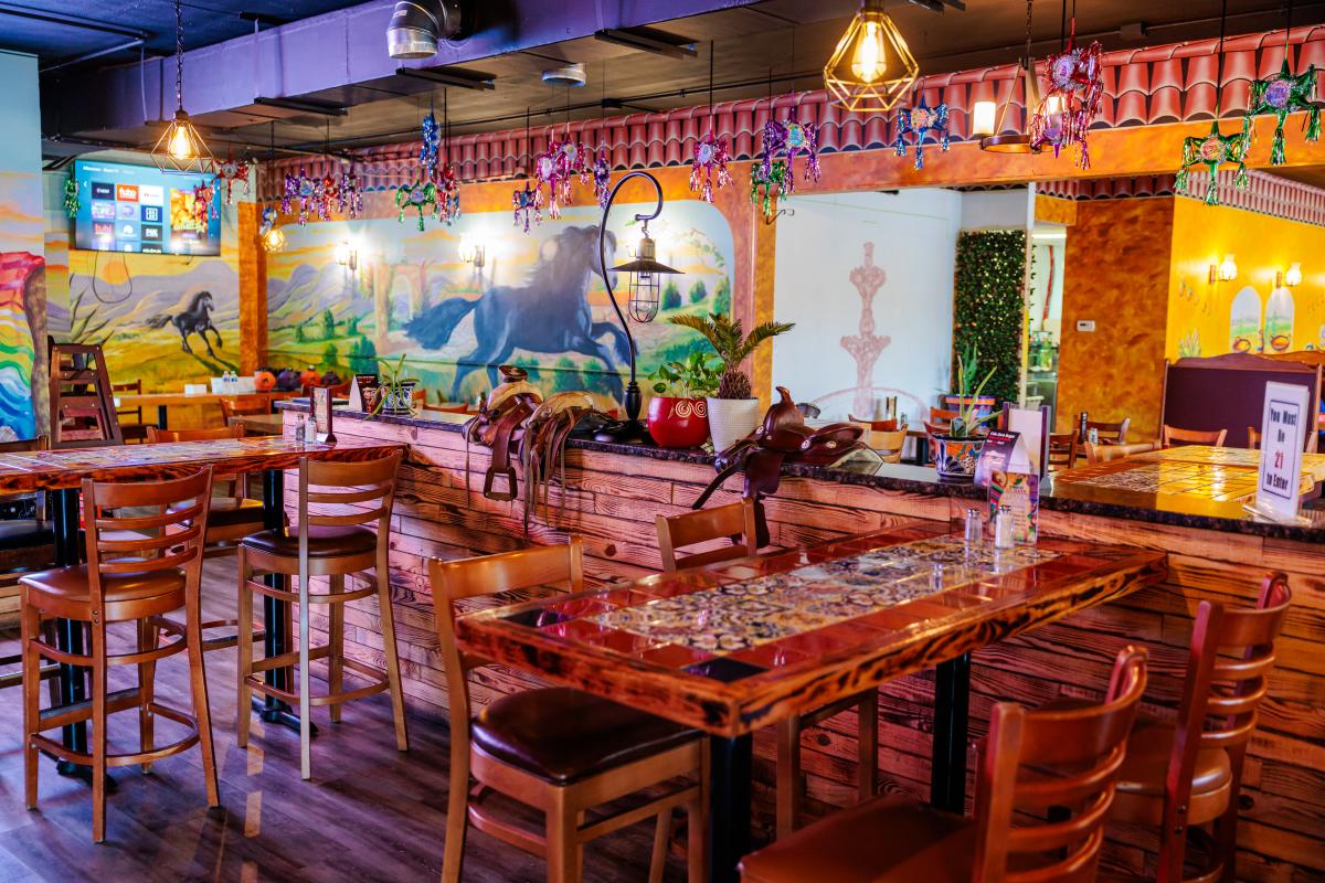 Bright and colorful interior of a restaurant decorated with vibrant murals depicting horses and landscapes on the walls. The space features wooden tables and chairs, with some tables adorned with intricate tile mosaics. Decorative lanterns hang from the ceiling, casting a warm glow. Small piñatas are suspended from above, adding a festive touch. A saddle is displayed on a wooden divider, enhancing the rustic and playful ambiance. Various potted plants and decor items are placed around, contributing to the lively atmosphere. A sign on one table indicates that patrons must be 21 or older to enter.