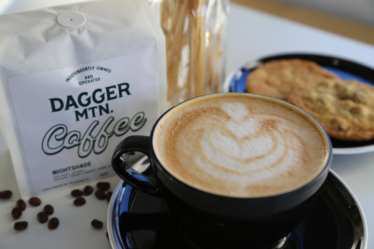 A latte in a dark mug on a saucer sits near a bag of coffee and a plate of cookies.