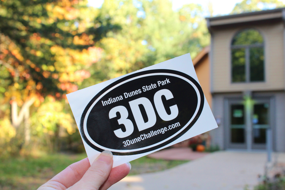A hand holds up a black oval sticker with white text that says 3DC and Indiana Dunes State Park