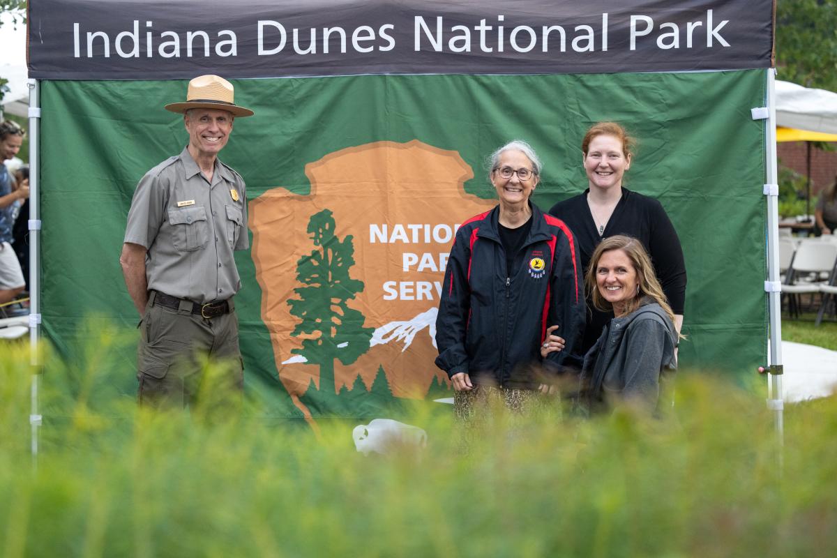 A group photo at Indiana Dunes National Park features a uniformed park ranger and three women standing in front of a National Park Service banner. The setting is an outdoor event with people, tents, and chairs visible in the background, while greenery in the foreground frames the scene.