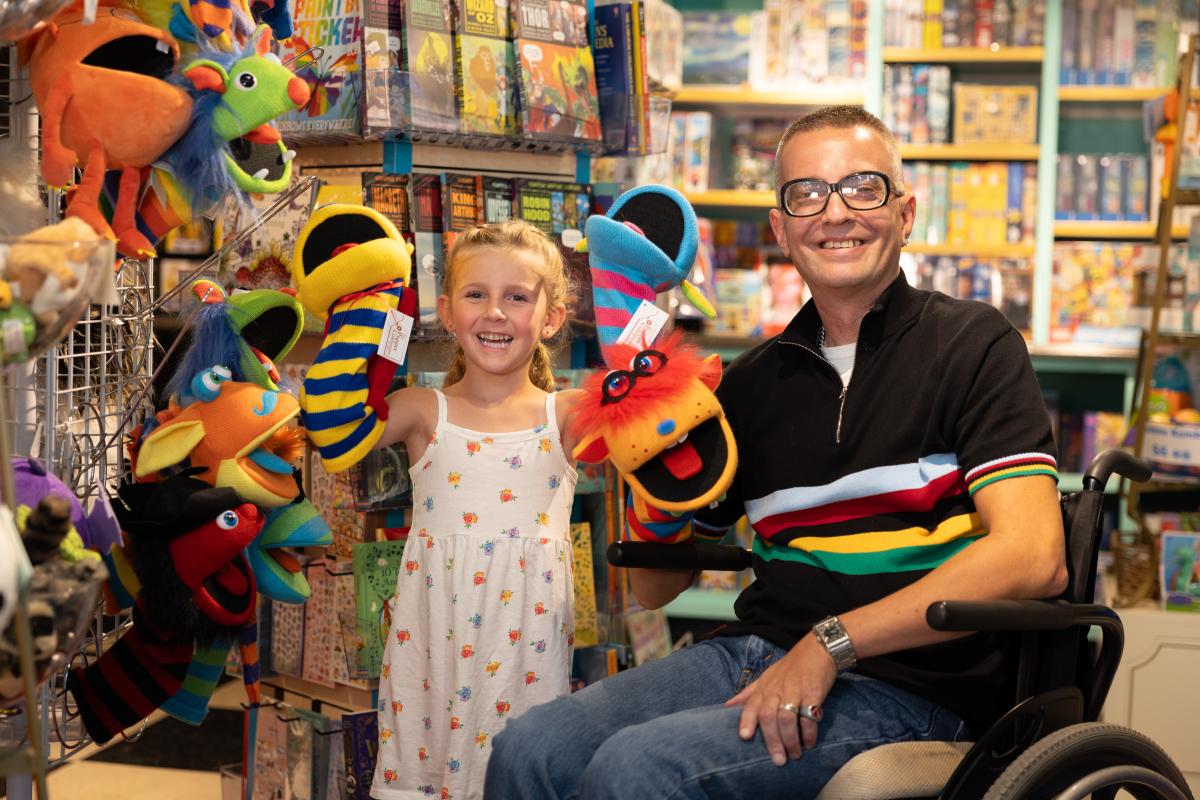 A young child stands next to a man sitting in a wheelchair. Both are smiling broadly and holding puppets.