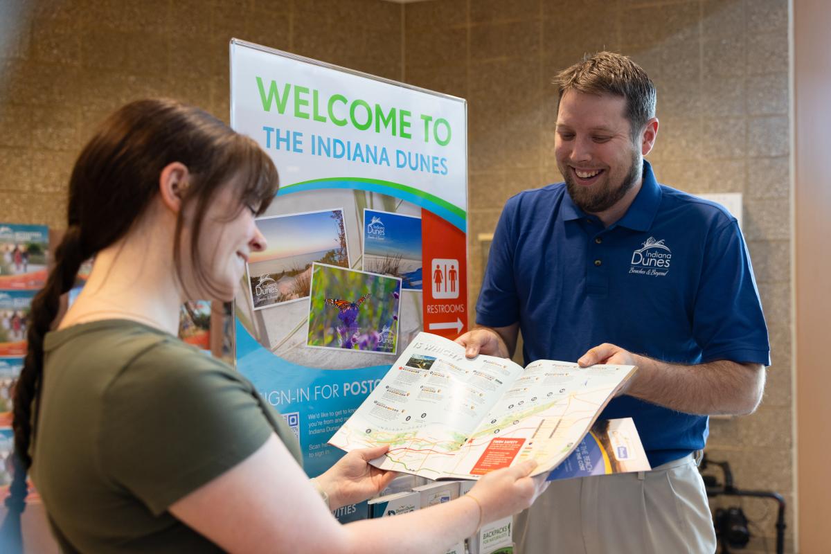 A man wearing a blue polo shows a brochure to a girl in a green shirt. They are standing next to a sign that says, "Welcome to the Indiana Dunes"